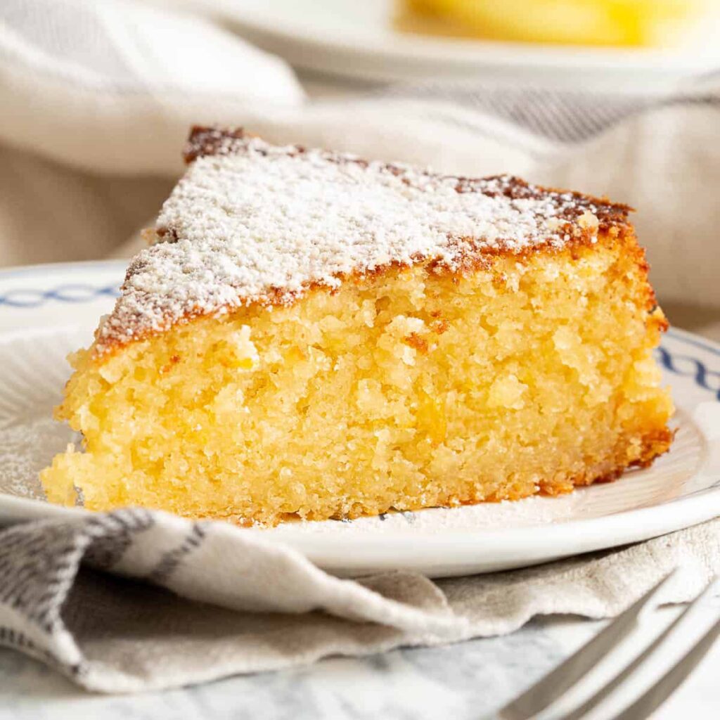 A golden Lemon Ricotta Cake sits on a white plate, adorned with a glossy lemon glaze drizzled over the top. A dusting of powdered sugar and delicate lemon zest curls decorate the surface, adding charm and elegance. The soft, moist crumb is visible through a slice, showcasing its delicate texture and vibrant citrus flavors.