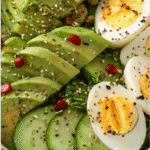 A colorful and nutritious Avocado Cucumber Egg Bowl Recipe with hard-boiled eggs, sliced avocado, cucumber rounds, sesame seeds, herbs, and olive oil served in a white ceramic bowl.
